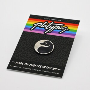 May include: A black and white enamel pin featuring a duck with a yin yang design. The pin is on a black card with rainbow stripes and the text "Platypus" in white. The card also says "Soft Enamel Pin" and "Made by Misfits in the UK".
