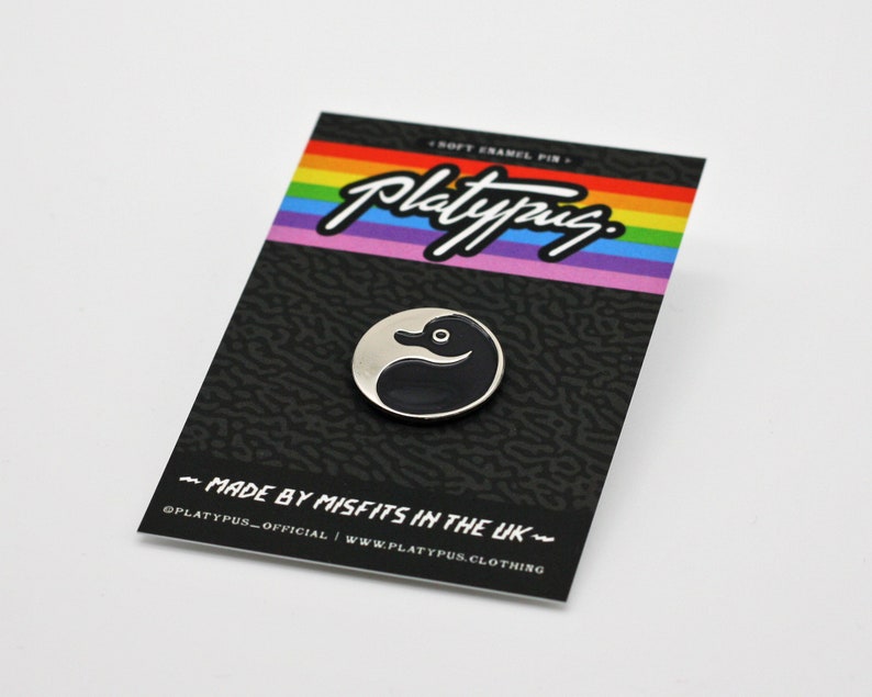 May include: A black and white enamel pin featuring a duck with a yin yang design. The pin is on a black card with rainbow stripes and the text "Platypus" in white. The card also says "Soft Enamel Pin" and "Made by Misfits in the UK".