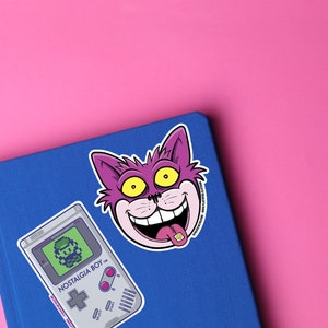 May include: A blue notebook with a purple cat sticker with a yellow eye and a pink tongue sticking out. The sticker says "Nostalgia Boy". There is also a sticker of a grey Game Boy with a pixelated character on the screen.