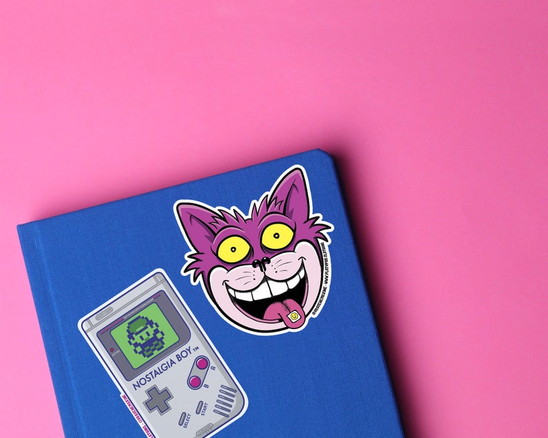 May include: A blue notebook with a purple cat sticker with a yellow eye and a pink tongue sticking out. The sticker says "Nostalgia Boy". There is also a sticker of a grey Game Boy with a pixelated character on the screen.