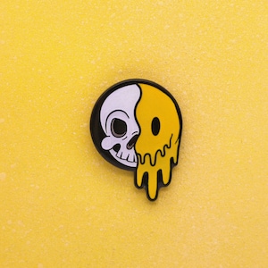 May include: A round, black and white pin with a skull on the left and a melting yellow smiley face on the right. The pin is set against a textured yellow background.