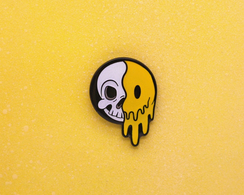 May include: A round, black and white pin with a skull on the left and a melting yellow smiley face on the right. The pin is set against a textured yellow background.