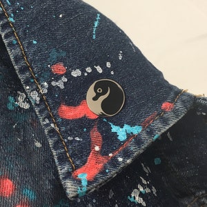 May include: A black and white yin yang symbol pin on a blue denim jacket with colorful paint splatters.