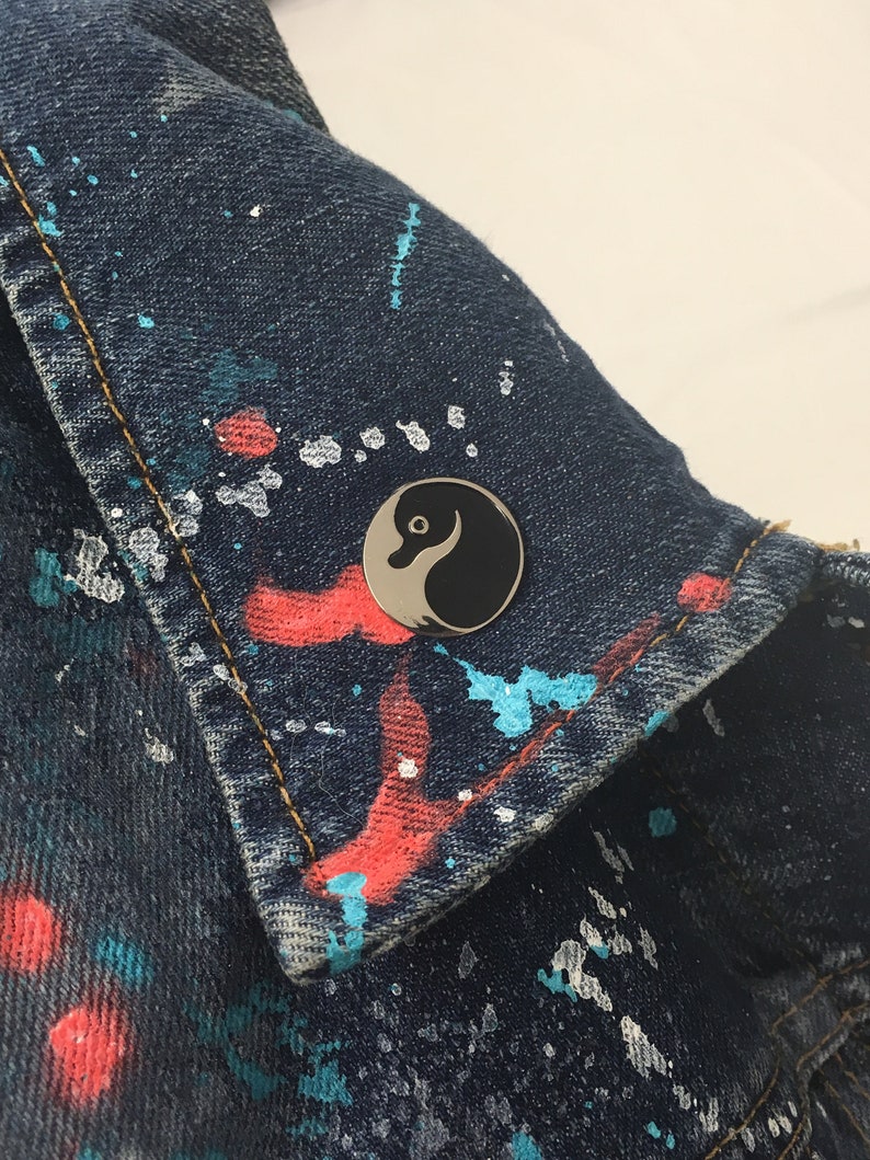 May include: A black and white yin yang symbol pin on a blue denim jacket with colorful paint splatters.