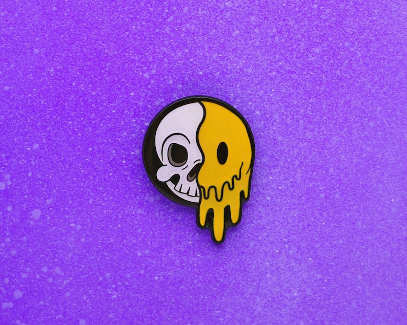 May include: A round enamel pin featuring a split design: a white skull on the left and a melting yellow smiley face on the right. The pin has a black border and is set against a purple background.