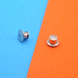 May include: Two silver-colored pin backs, one with a post and the other with a cap, are displayed against a blue and orange background. The pin backs are round with a flat base and a cylindrical post.