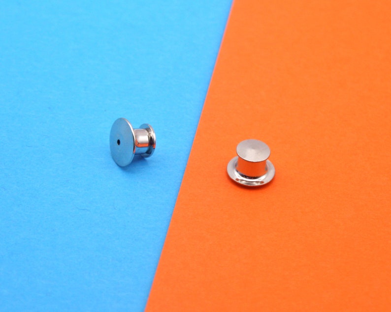 May include: Two silver-colored pin backs, one with a post and the other with a cap, are displayed against a blue and orange background. The pin backs are round with a flat base and a cylindrical post.