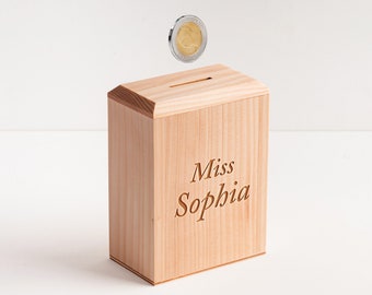 May include: A wooden piggy bank with a slot on top. The piggy bank is light brown and has the name "Miss Sophia" engraved on the front.