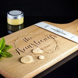 May include: A personalized wooden cutting board with the text "The Armstrongs Est. 2019" engraved on it. The board is made of light wood and has a handle. There is a small jar of balm and a sprig of mint on the board.