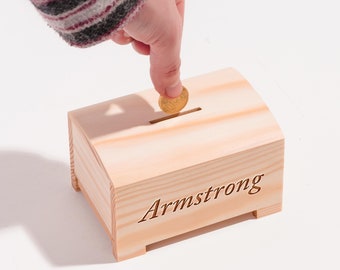 May include: A wooden piggy bank with the name "Armstrong" engraved on the front. A hand is placing a gold coin in the slot.