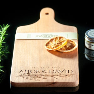 K&ouml;nnte beinhalten: Ein Holzbrett zum Schneiden mit Griff, personalisiert mit dem Text "EST. 13-11-2020 Alice & David". Das Brett hat zwei getrocknete Orangenscheiben und ein kleines Glas mit Holzpflegemittel mit der Aufschrift "CYANWOOD" darauf.