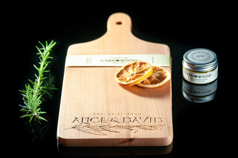 K&ouml;nnte beinhalten: Ein Holzbrett zum Schneiden mit Griff, personalisiert mit dem Text "EST. 13-11-2020 Alice & David". Das Brett hat zwei getrocknete Orangenscheiben und ein kleines Glas mit Holzpflegemittel mit der Aufschrift "CYANWOOD" darauf.