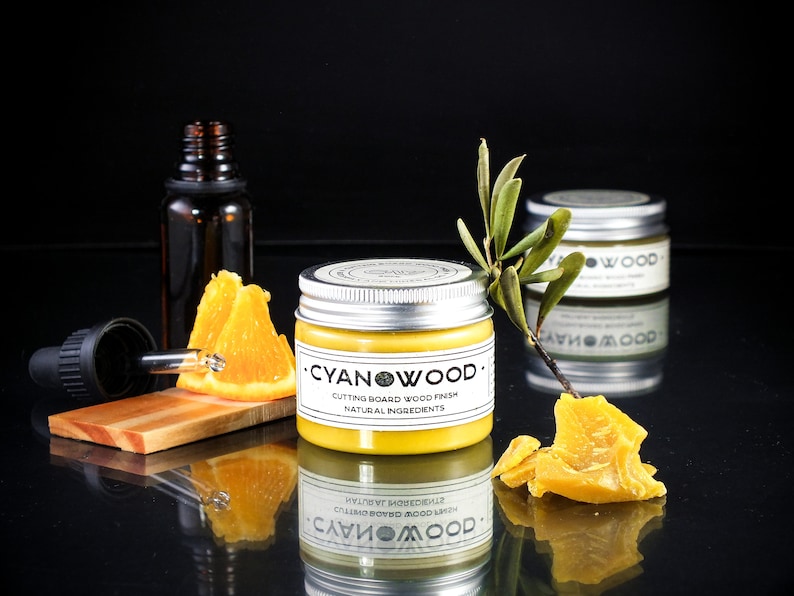 K&ouml;nnte beinhalten: Ein kleines Glas mit gelber Holzpolitur und einem silbernen Deckel. Das Glas ist mit "CYANWOOD - CUTTING BOARD WOOD FINISH - NATURAL INGREDIENTS" beschriftet. Im Hintergrund sind Orangenscheiben und eine Tropfflasche zu sehen.