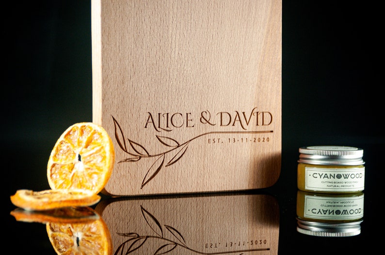 May include: A wooden chopping board with the text "Alice & David Est. 13-11-2020" engraved on it. A small jar of wood conditioner is in the foreground.