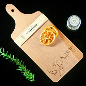 May include: A wooden chopping board with a handle, personalised with the text "Alice & David Est. 2020". The board is made by Cyanwood and has a natural wood finish. There are dried orange slices on the board.