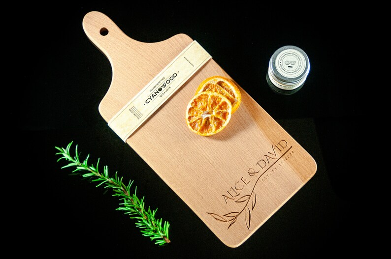 May include: A wooden chopping board with a handle, personalised with the text "Alice & David Est. 2020". The board is made by Cyanwood and has a natural wood finish. There are dried orange slices on the board.
