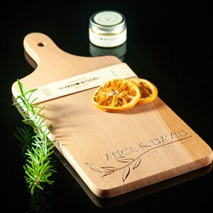 May include: A personalised wooden chopping board with the text "Alice & David" engraved on it. The board is light brown and has a handle. There are two dried orange slices and a sprig of rosemary on the board. The board is sitting on a black surface.