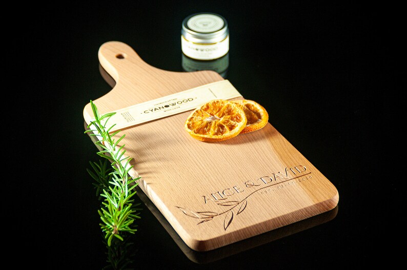 May include: A personalised wooden chopping board with the text "Alice & David" engraved on it. The board is light brown and has a handle. There are two dried orange slices and a sprig of rosemary on the board. The board is sitting on a black surface.
