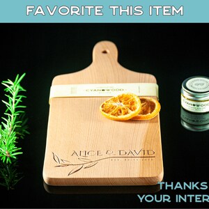 May include: A personalised wooden chopping board with the text "Alice & David Est. 2020" engraved on it. The board is light brown and has a handle. Two dried orange slices are on the board. The board is on a black surface.