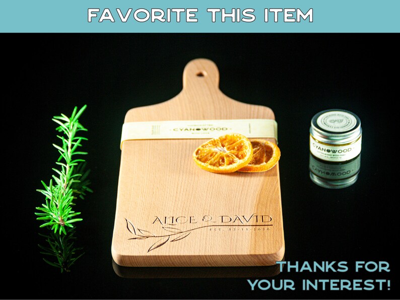 May include: A personalised wooden chopping board with the text "Alice & David Est. 2020" engraved on it. The board is light brown and has a handle. Two dried orange slices are on the board. The board is on a black surface.
