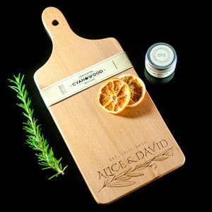 K&ouml;nnte beinhalten: Ein Schneidebrett aus Holz mit einem Griff, graviert mit dem Text "EST. 13-11-2020 Alice & David". Das Brett ist auch mit "Handcrafted Cyanwood. With Love." beschriftet.