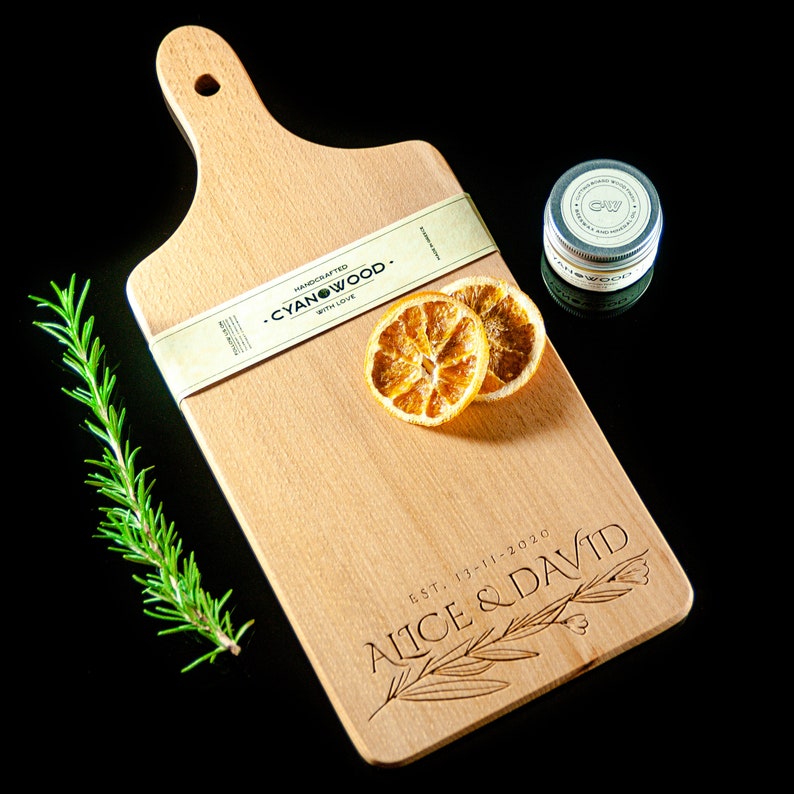 K&ouml;nnte beinhalten: Ein Schneidebrett aus Holz mit einem Griff, graviert mit dem Text "EST. 13-11-2020 Alice & David". Das Brett ist auch mit "Handcrafted Cyanwood. With Love." beschriftet.