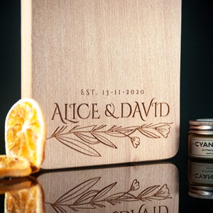 K&ouml;nnte beinhalten: Ein Schneidebrett aus Holz mit dem eingravierten Text "EST. 13-11-2020 ALICE & DAVID". Das Brett ist mit einem floralen Design verziert und hat eine nat&uuml;rliche Holzoptik.
