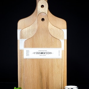 K&ouml;nnte beinhalten: Drei Holz-Schneidebretter mit Griffen, die &uuml;bereinander gestapelt sind. Die Bretter sind aus hellem Holz und haben eine nat&uuml;rliche Maserung. Das oberste Brett hat ein wei&szlig;es Etikett mit dem aufgedruckten Text "HANDCRAFTED - CYANWOOD - WITH LOVE". Ein kleines Honigglas steht auf einer schwarzen Oberfl&auml;che vor den Brettern.