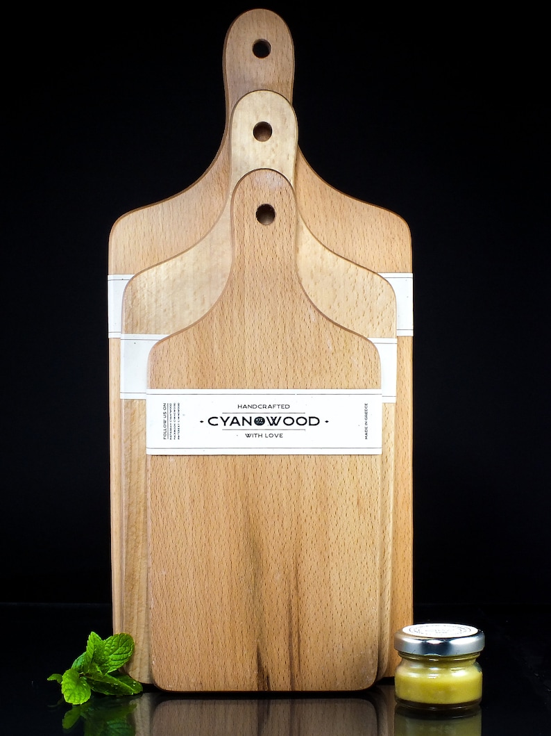 K&ouml;nnte beinhalten: Drei Holz-Schneidebretter mit Griffen, die &uuml;bereinander gestapelt sind. Die Bretter sind aus hellem Holz und haben eine nat&uuml;rliche Maserung. Das oberste Brett hat ein wei&szlig;es Etikett mit dem aufgedruckten Text "HANDCRAFTED - CYANWOOD - WITH LOVE". Ein kleines Honigglas steht auf einer schwarzen Oberfl&auml;che vor den Brettern.