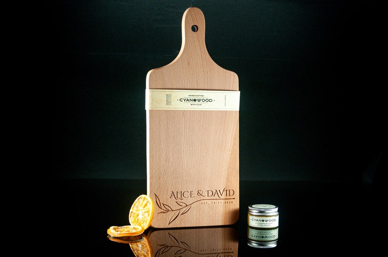 May include: A personalised wooden chopping board with a handle, engraved with the text "Alice & David Est. 12-11-2020" and a leafy design. The board is made by Cyanwood.