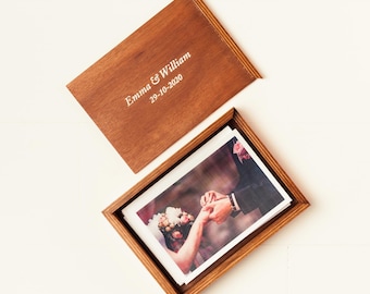 May include: A wooden photo box with a hinged lid, containing printed photographs. The lid is engraved with the names "Emma & William" and the date "29-10-2020". The box is made of dark brown wood.