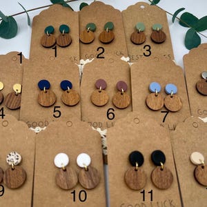 May include: A collection of wooden stud earrings with various colored tops, including green, blue, and gold. The earrings are displayed on brown tags with the words "GOOD LUCK" and numbers. Some earrings have a circular wooden base and a smaller, contrasting top.