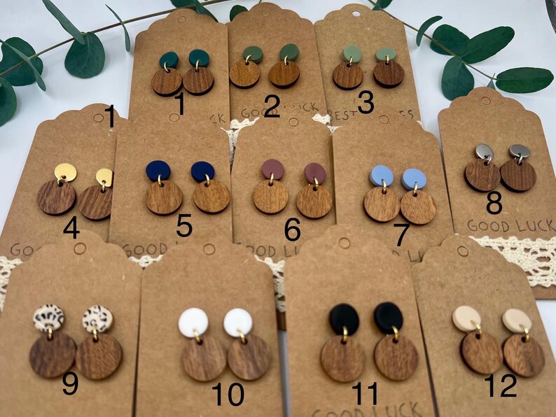 May include: A collection of wooden stud earrings with various colored tops, including green, blue, and gold. The earrings are displayed on brown tags with the words "GOOD LUCK" and numbers. Some earrings have a circular wooden base and a smaller, contrasting top.