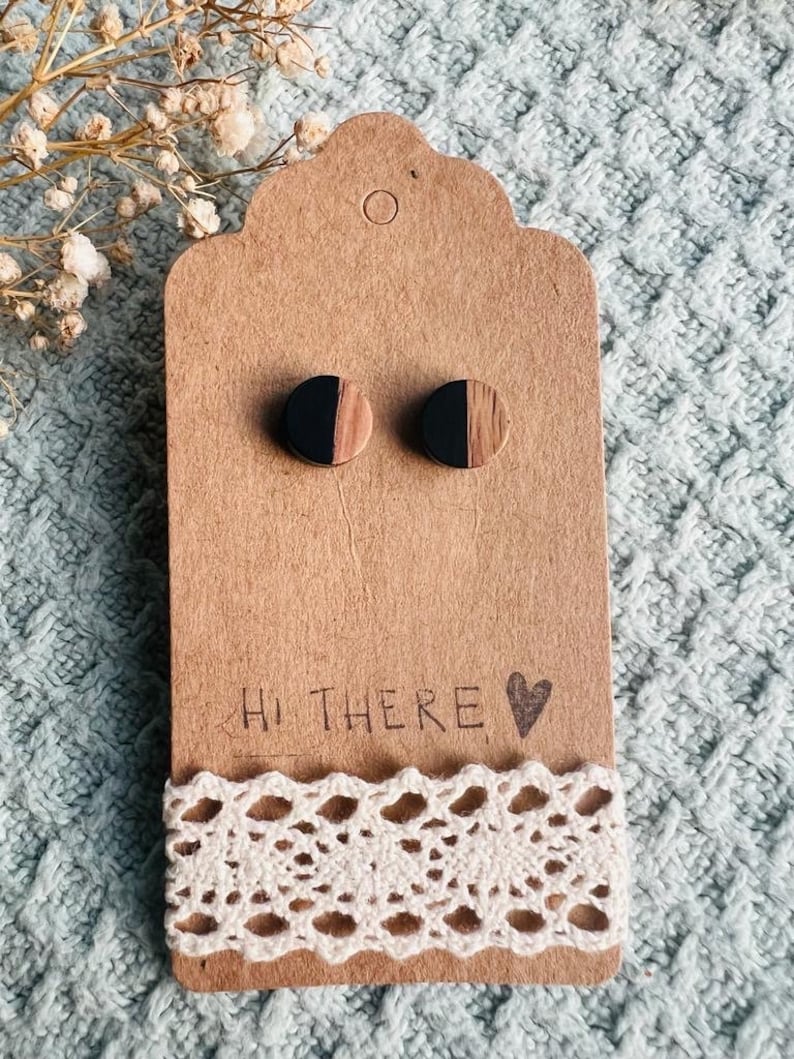 May include: A pair of wooden earrings with a black and wood grain design. The earrings are displayed on a brown card with the text "Hi There" and a heart.