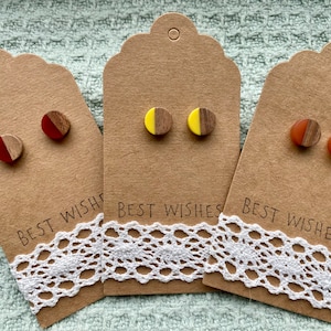May include: Three pairs of wooden stud earrings with a half-circle of colored resin on the front. The earrings are displayed on brown cards with the text "Best Wishes".