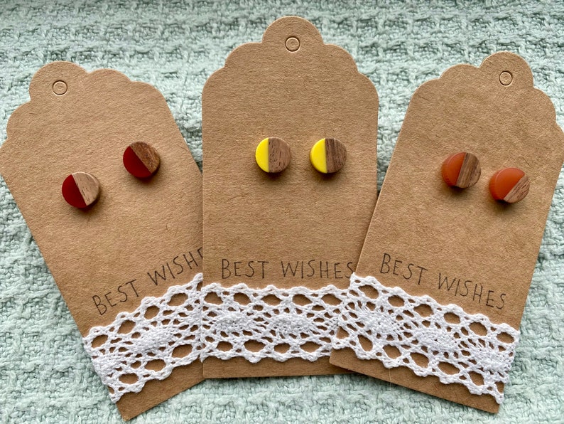 May include: Three pairs of wooden stud earrings with a half-circle of colored resin on the front. The earrings are displayed on brown cards with the text "Best Wishes".