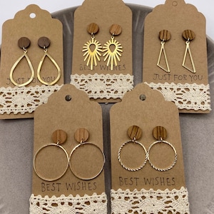 Könnte beinhalten: Fünf Paar goldene Ohrringe auf braunem Karton mit dem Text "Beste Wünsche" und "Nur für dich". Die Ohrringe werden auf einem weißen Spitzenhintergrund präsentiert.