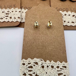 May include: A display of earrings on brown cardboard tags with cream-colored lace trim. The earrings include small gold studs and larger wooden disc earrings with colored tops. The text "GOOD LUCK" is partially visible on the tags.