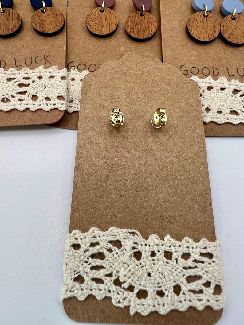 May include: A display of earrings on brown cardboard tags with cream-colored lace trim. The earrings include small gold studs and larger wooden disc earrings with colored tops. The text "GOOD LUCK" is partially visible on the tags.