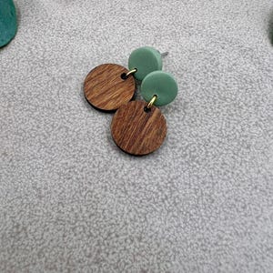 May include: A pair of earrings featuring a combination of wood and green elements. The earrings have a wooden, circular design with a natural wood grain pattern, paired with two green, circular studs. The earrings are connected with gold-colored hardware.