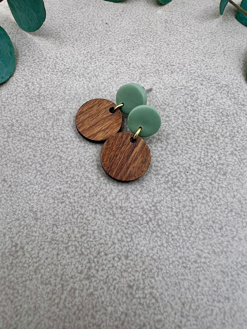 May include: A pair of earrings featuring a combination of wood and green elements. The earrings have a wooden, circular design with a natural wood grain pattern, paired with two green, circular studs. The earrings are connected with gold-colored hardware.
