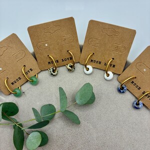 Könnte beinhalten: Goldene Creolen-Ohrringe mit verschiedenen Perlenmustern: grün, gesprenkelt, weiß und blau. Jeder Ohrring wird auf einer braunen Karte mit dem Schriftzug "MUCH LOVE" präsentiert. Ein Eukalyptuszweig liegt im Vordergrund.