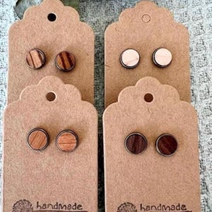 Könnte beinhalten: Vier Paar handgefertigte Holzohrringe, die auf braunem Karton mit dem aufgedruckten Wort "handgefertigt" präsentiert werden. Die Ohrringe sind in verschiedenen Holztönen und Mustern gehalten.
