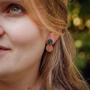 May include: A pair of earrings featuring a small, dark green circle above a larger, round wooden disc. The earrings are worn on a person's ear, with the focus on the jewelry. The earrings are a simple, minimalist design.
