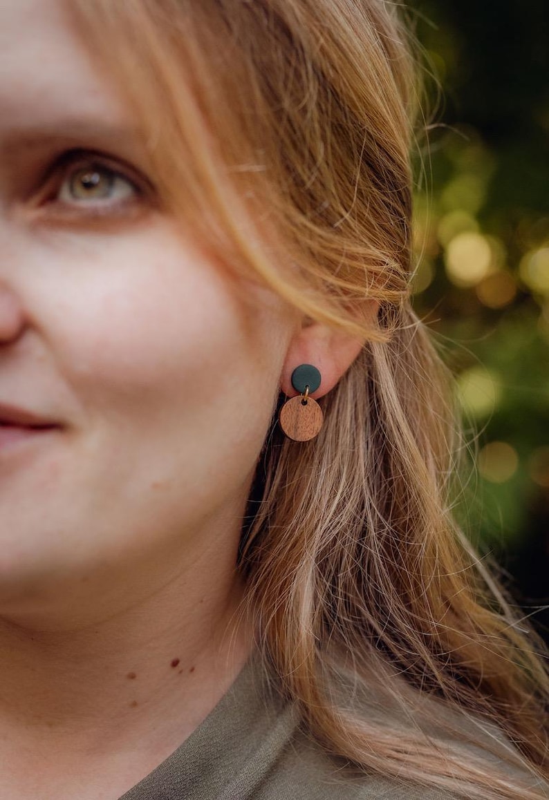 May include: A pair of earrings featuring a small, dark green circle above a larger, round wooden disc. The earrings are worn on a person's ear, with the focus on the jewelry. The earrings are a simple, minimalist design.