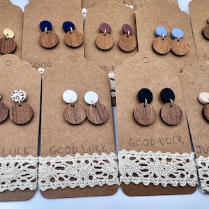 May include: A collection of wooden earrings with various colored tops, including gold, blue, black, and white. The earrings are displayed on brown tags with the words "GOOD LUCK" and "JUST FOR YOU" printed on them, with a lace trim.