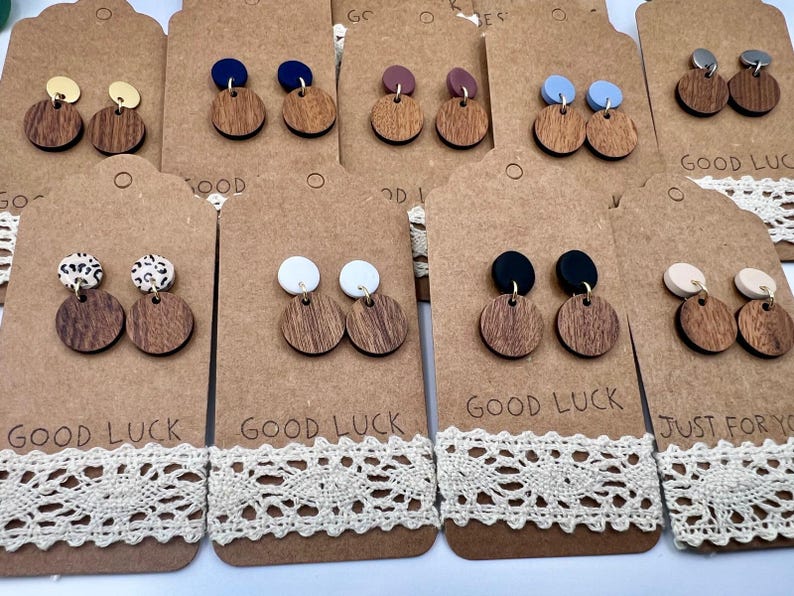 May include: A collection of wooden earrings with various colored tops, including gold, blue, black, and white. The earrings are displayed on brown tags with the words "GOOD LUCK" and "JUST FOR YOU" printed on them, with a lace trim.