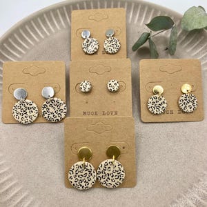 Könnte beinhalten: Drei Paar Ohrringe mit Leopardenmuster auf Präsentationskarten. Die Ohrringe sind aus Polymer Clay gefertigt und haben goldene und silberne Akzente. Die Karten tragen den Aufdruck "Much Love".