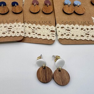 May include: Wooden earrings with a white top and a brown, circular bottom. The earrings are displayed on a brown card with the words "GOOD LUCK" printed on it. The earrings have gold-colored posts.