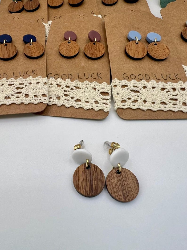 May include: Wooden earrings with a white top and a brown, circular bottom. The earrings are displayed on a brown card with the words "GOOD LUCK" printed on it. The earrings have gold-colored posts.